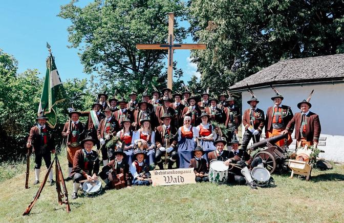 Das Bild zeigt eine große Gruppe von Menschen in traditioneller Tracht einer Schützenkompanie, die vor einem Kreuz und einer kleinen Kapelle im Freien für ein Gruppenfoto posieren.