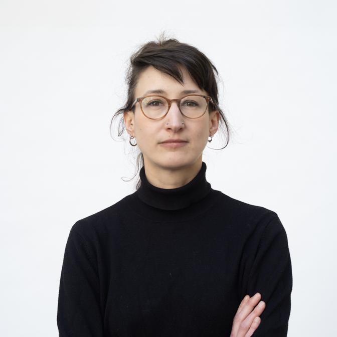 Frau mit Brille und schwarzen Rollkragenpullover, verschränkte Arme, vor weißem Hintergrund.