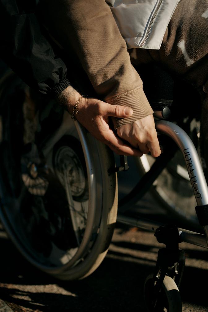 Zwei Menschen halten sich an den Händen, einer sitzt im Rollstuhl. Das Sonnenlicht wirft Schatten auf das Rad und ihre Hände und vermittelt so Unterstützung und Verbundenheit.