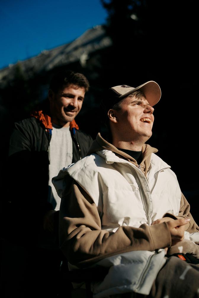 Ein lächelnder Mann im Rollstuhl, der eine Mütze und eine Weste trägt, sitzt draußen unter strahlend blauem Himmel, während ein anderer Mann hinter ihm steht.