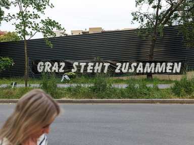 Eine Frau geht an einer Mauer vorbei, an der ein Banner mit der Aufschrift „Graz steht zusammen“ hängt, umgeben von Grünflächen und Bäumen.
