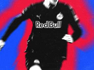 Eine verschwommene Gestalt in einem schwarzen Fußballtrikot mit einem „Red Bull“-Logo rennt vor einem leuchtend roten und blauen Hintergrund.