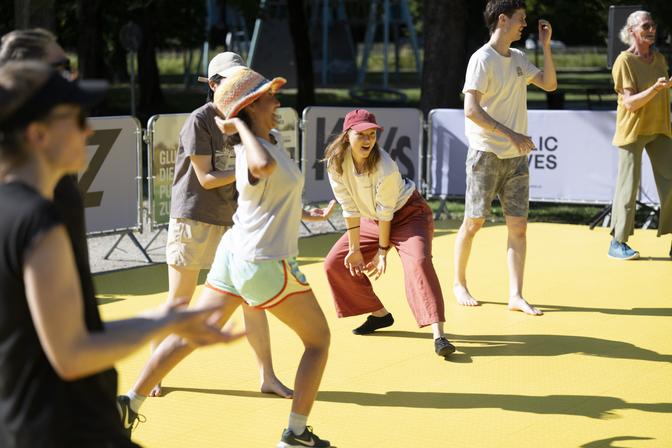 Menschen tanzen energisch an einem sonnigen Tag in legerer Sommerkleidung auf einer gelben Matte im Freien im Rahmen von Impulstanz.