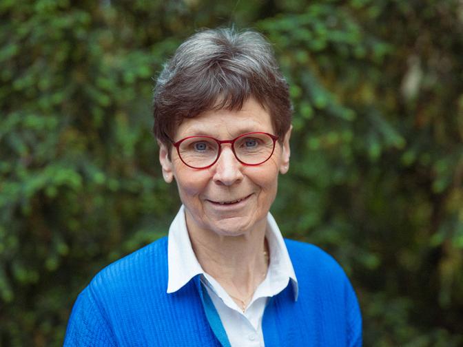 Ältere Frau mit kurzen Haaren und roter Brille im blauen Pullover, lächelnd vor einem verschwommenen Hintergrund mit grünen Blättern.