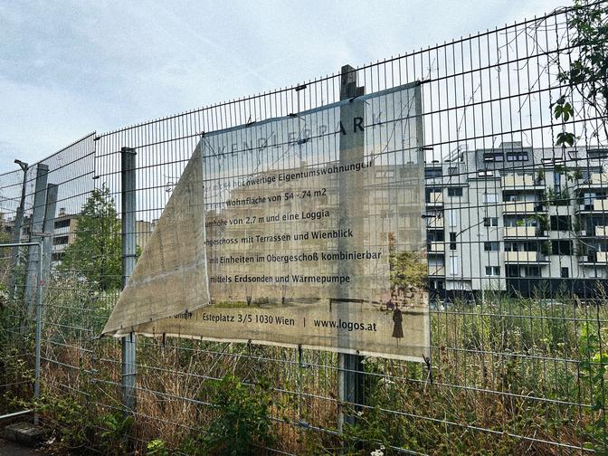 Eine zerrissene Werbung an einem Drahtzaun mit überwuchertem Gras, in der Nähe eines Wohnhauses und eines leeren Grundstücks.