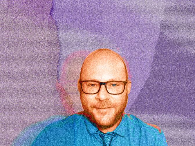 Eine Person mit Brille und Bart trägt ein blaues Hemd und eine Krawatte, vor einem farbenfrohen, abstrakten Hintergrund.