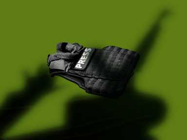 Eine schwarze Presseweste mit der Aufschrift „PRESS“ liegt auf einem grünen Hintergrund und wirft einen deutlichen Schatten.