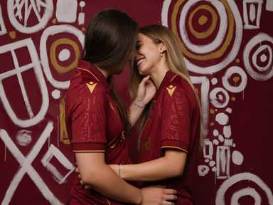 Zwei Frauen in roten FK Austria Wien Trikots lächeln sich an und umarmen sich vor einer stilisierten Wand mit weißen und goldenen Kreisen und Mustern.