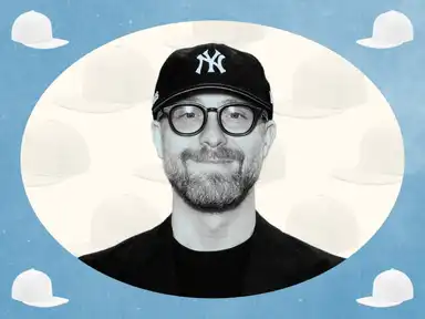 Ein Mann mit Brille und Bart, trägt eine schwarze Mütze mit einem "NY"-Logo, vor einem Hintergrund mit weißen Mützenabbildungen.