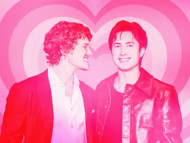 Die Hauptdarsteller der Serie "Heated Rivalry" in Anzügen lächeln vor einem rosa Hintergrund mit Herzmuster.