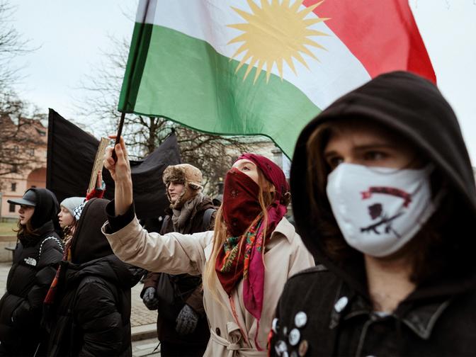 Demonstranten mit Masken und Winterkleidung halten an einem bewölkten Tag im Freien kurdische Flaggen und schwarze Banner hoch.