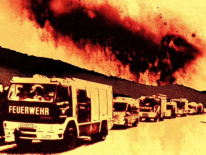 Feuerwehrfahrzeuge stehen auf einer Straße vor einem großen, dunklen Rauch- und Flammenhimmel während eines Feuers.