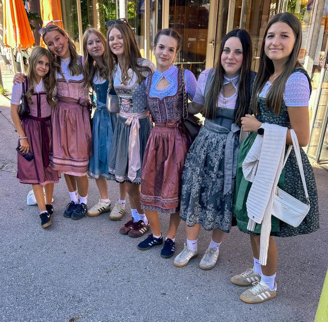 Gruppe von sieben Mädchen in bunten Dirndln und Sportschuhen, die draußen nebeneinander stehen und lächeln.