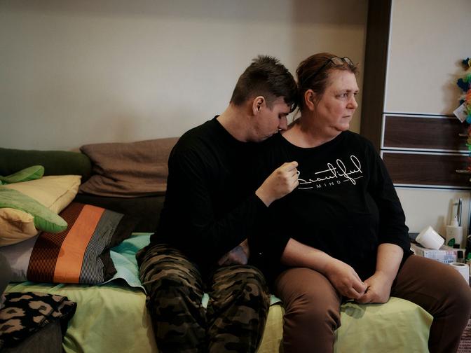 Eine Person sitzt dicht neben einer anderen auf einem Bett und lehnt sich an deren Schulter. Der Raum ist mit bunten Kissen und Dekorationen ausgestattet.