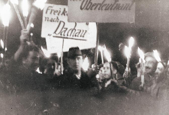 Historisches Schwarz-Weiß-Foto einer nächtlichen Prozession, auf dem Menschen Fackeln und Schilder mit deutschen Aufschriften tragen.