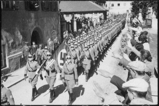 Während einer Parade in historischen Kostümen marschieren Nazi-Soldaten in Formation eine Straße entlang, flankiert von Zuschauern, die den Hitlergruß zeigen.