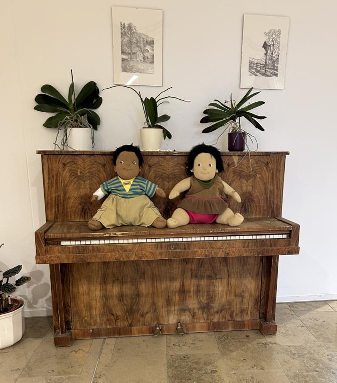 Zwei Plüschpuppen sitzen auf einem hölzernen Klavier, über dem Topfpflanzen und gerahmte Bilder stehen, auf einem Fliesenboden.