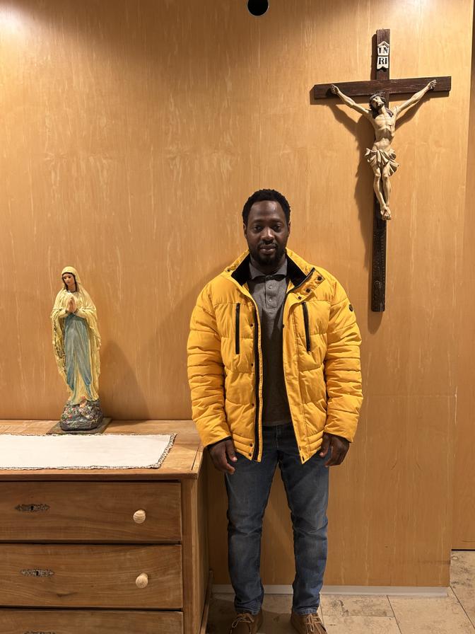 Ein Mann in einer gelben Jacke steht zwischen einer Statue der Jungfrau Maria auf einem Tisch und einem an der Wand befestigten Kruzifix.