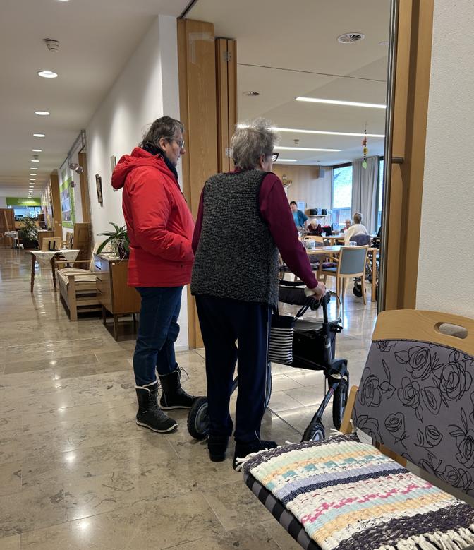 Eine Frau hilft einer älteren Frau mit einer Gehhilfe in einem gut beleuchteten Flur, im Hintergrund ist ein Gemeinschaftsraum zu sehen.