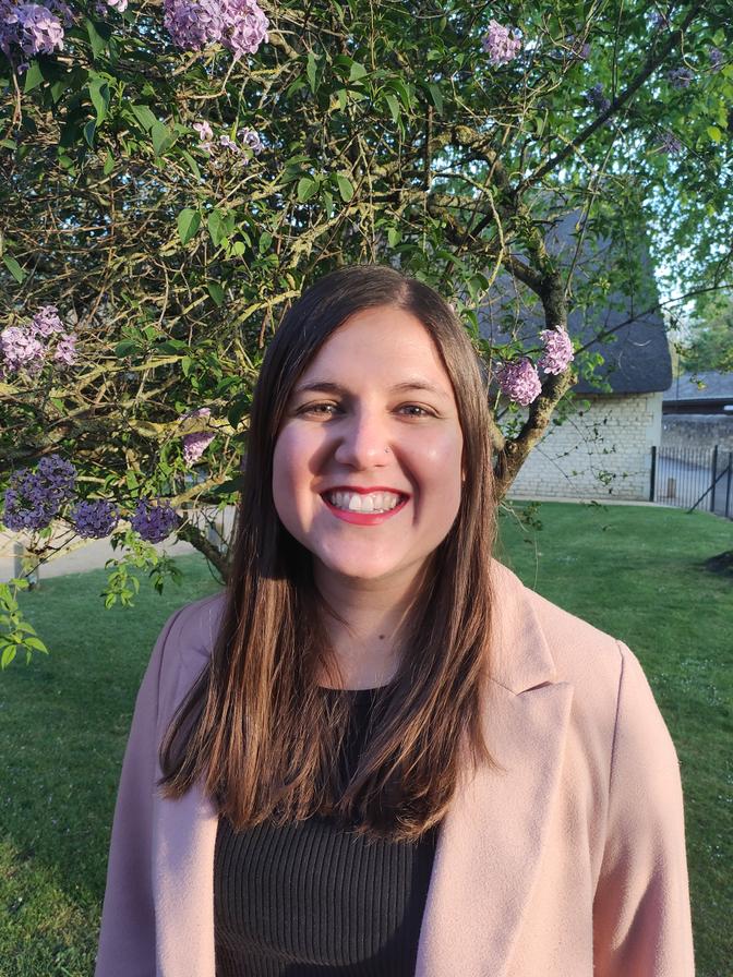Smiling woman with long brown hair in a pink coat and black top standing by a lilac-blooming tree in a sunny garden.
