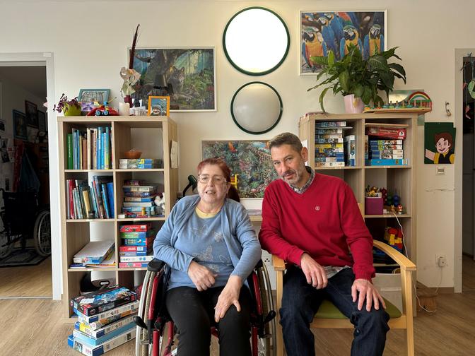 Eine Bewohnerin im Rollstuhl sitzt neben einem Mitarbeiter der Lebenshilfe im Wohnhaus; im Hintergrund stehen Bücherregale, Spiele und Pflanzen.
