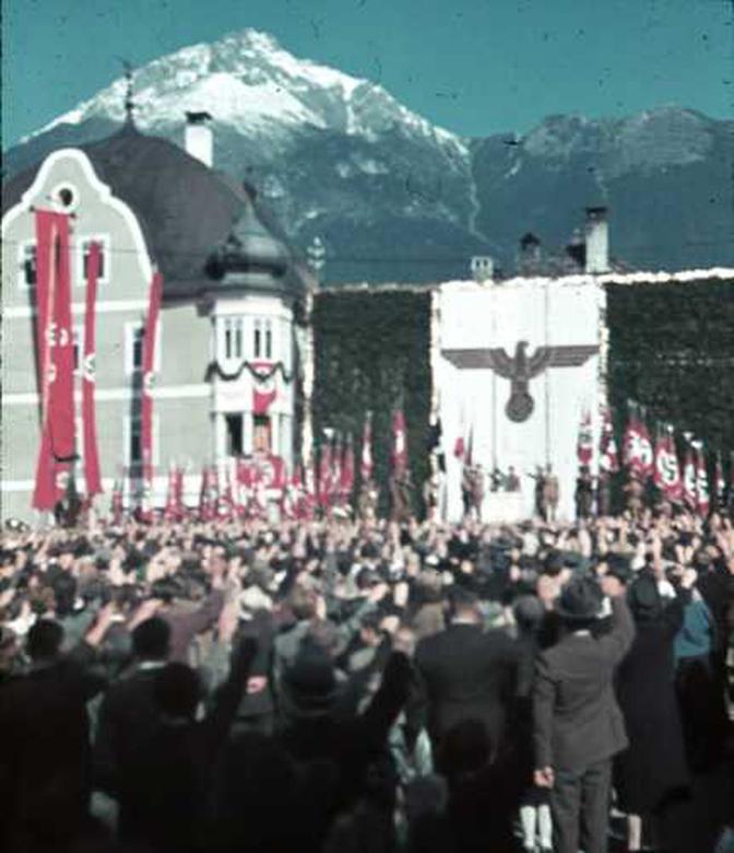 Bei einer Nazi-Kundgebung vor einem geschmückten Gebäude mit einem Hakenkreuzbanner hebt eine Menschenmenge die Hände zum Gruß. Im Hintergrund sind schneebedeckte Berge zu sehen.