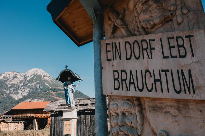 Eine Holzschnitzerei auf der steht "Ein Dorf lebt Brauchtum", dahinter eine Jesusstatue, Bauernhäuser und Berge.