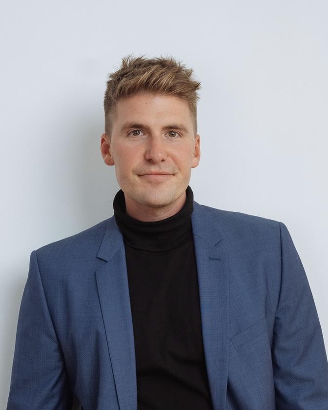 Ein Mann mit blonden Haaren trägt einen blauen Anzug und schwarzen Rollkragenpullover, vor einem hellen Hintergrund.