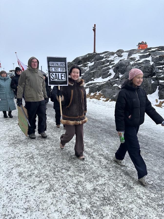 Menschen gehen auf einem verschneiten Weg, einer hält ein Schild mit der Aufschrift „Grönland ist nicht zu verkaufen!“ Im Hintergrund eine felsige Landschaft.